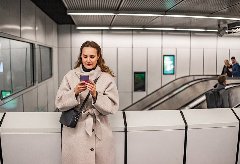 With a crossbody bag and a cozy coat, a woman scrolling through her phone, tuning out the rush of the metro station around her